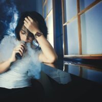 Das Bild zeigt eine Frau die an ein Fenster angelehnt ist und eine E-Zigarette raucht. Foto von Dede Avez von Pexels