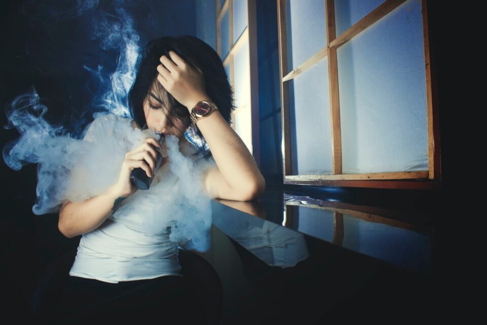 Das Bild zeigt eine Frau die an ein Fenster angelehnt ist und eine E-Zigarette raucht.  Foto von Dede Avez von Pexels