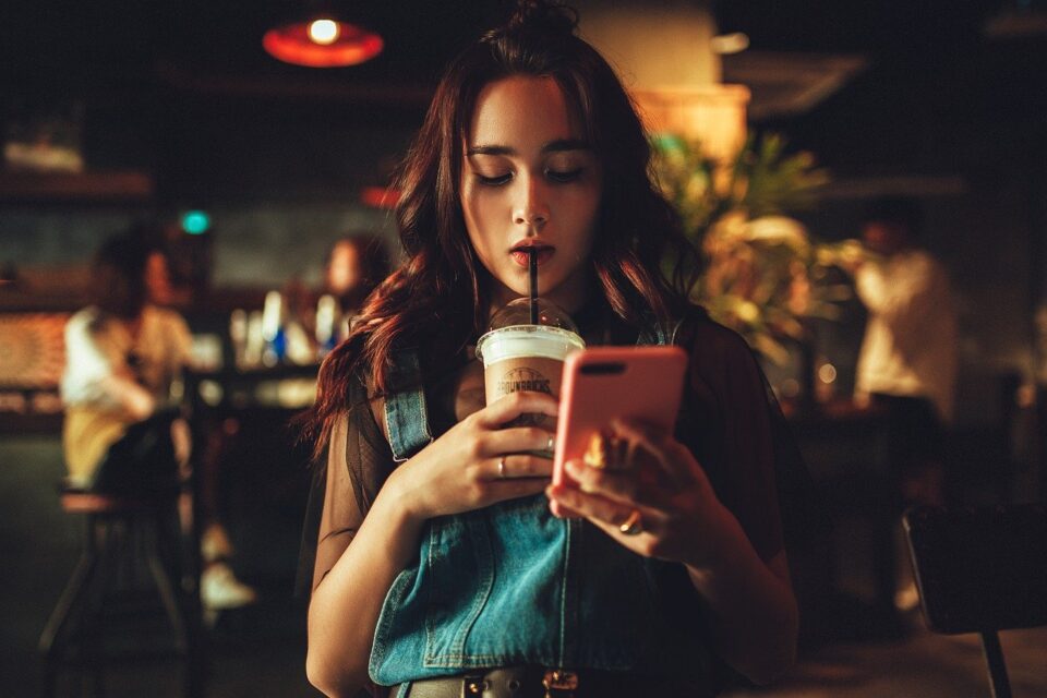 Das Foto zeigt eine junge Frau, die Kaffee schlürfend auf Ihr Smartphone schaut. Foto: Pexels.com by Averyanovphoto