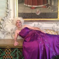 Drag Queen Edda liegt in einem pompösen lilafarbenem Kleid vor einer barocken Kulisse. Foto: SCHMITZ e.V.