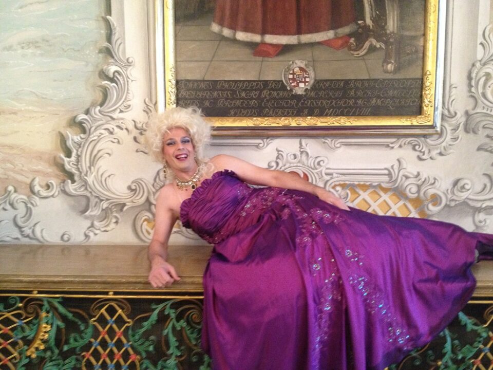 Drag Queen Edda liegt in einem pompösen lilafarbenem Kleid vor einer barocken Kulisse. Foto: SCHMITZ e.V. 
